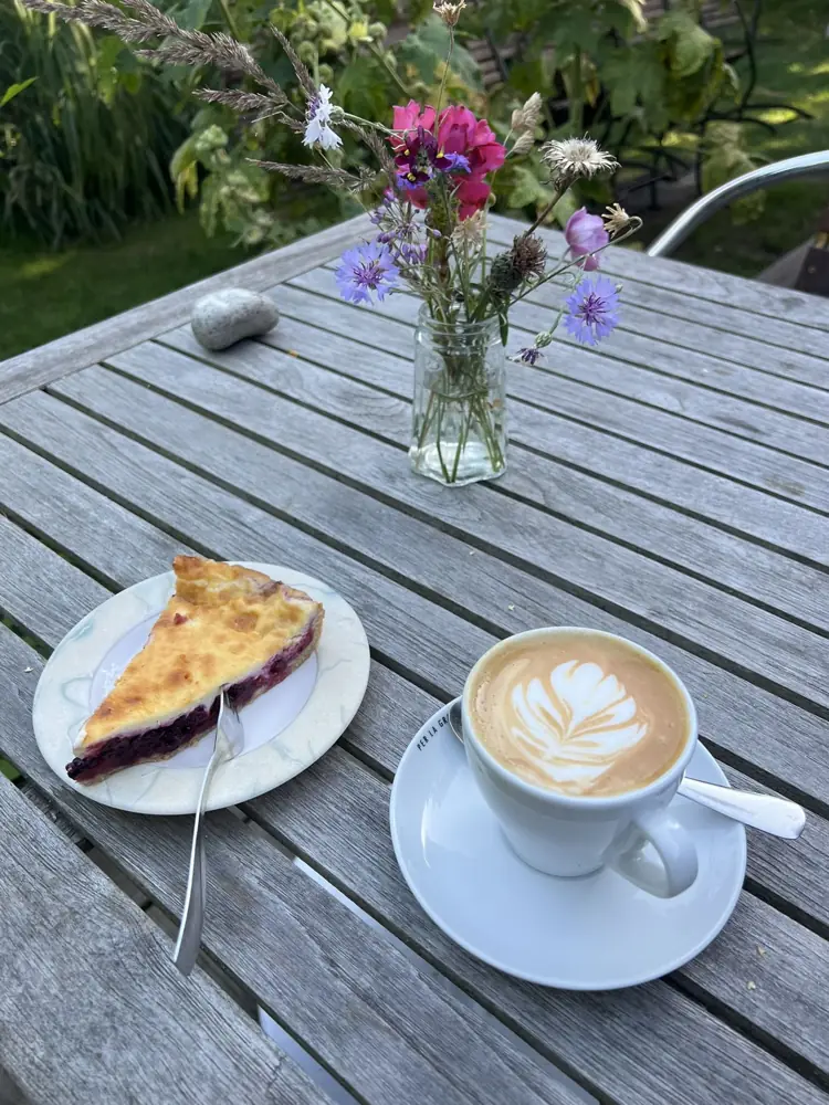 Ein Stück Beerenkuchen und ein Cappuccino auf dem Holztisch, daneben Wiesenblumen